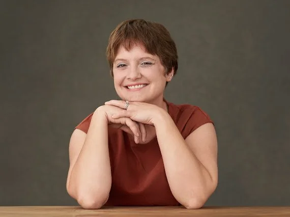 A woman with short brown hair smiling and resting her chin on her folded hands, sitting at a wooden table against a neutral background.