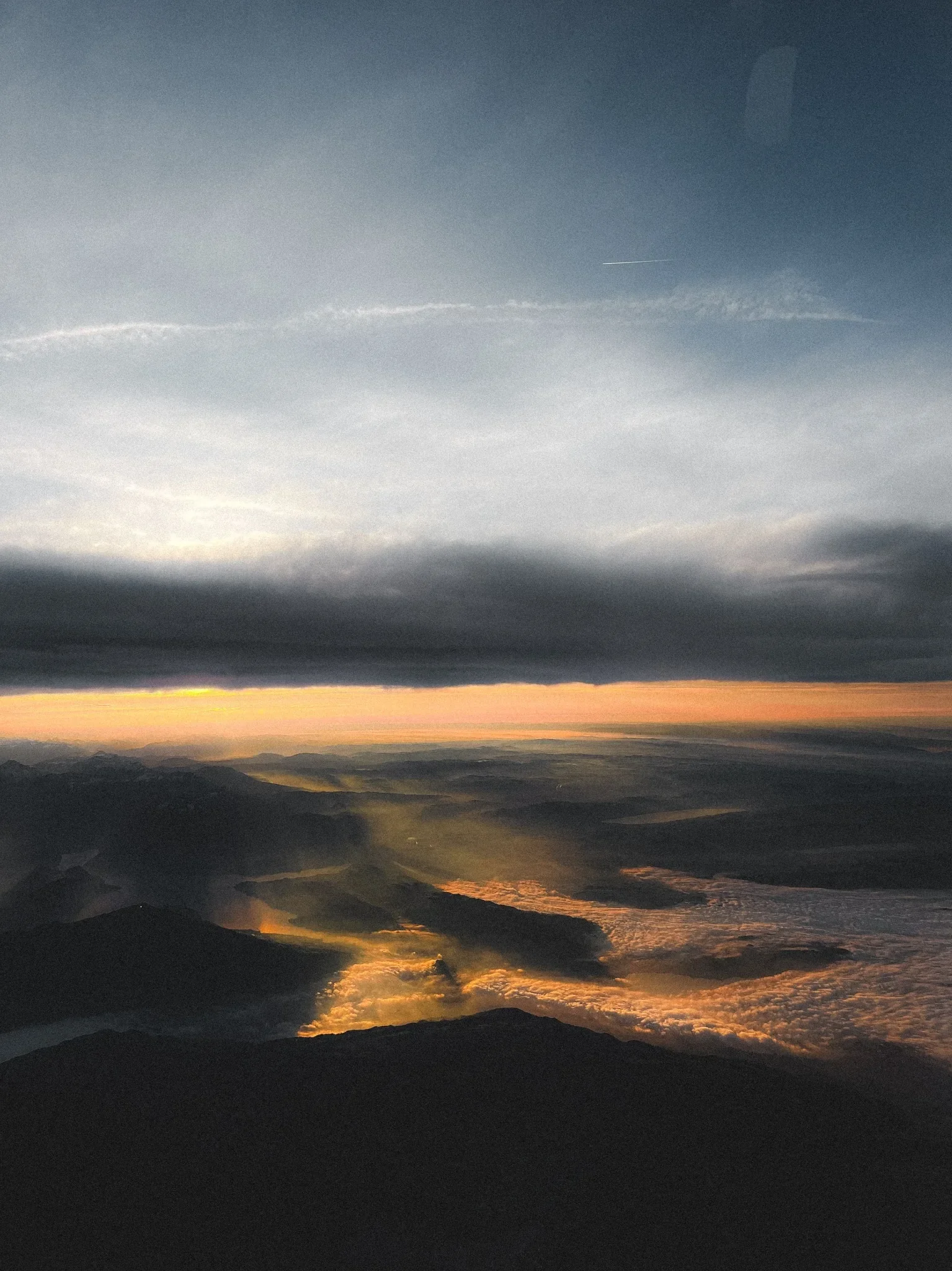 Sunset or sunrise over a mountainous landscape with clouds and the ocean, featuring dark foreground mountains and a bright horizon
