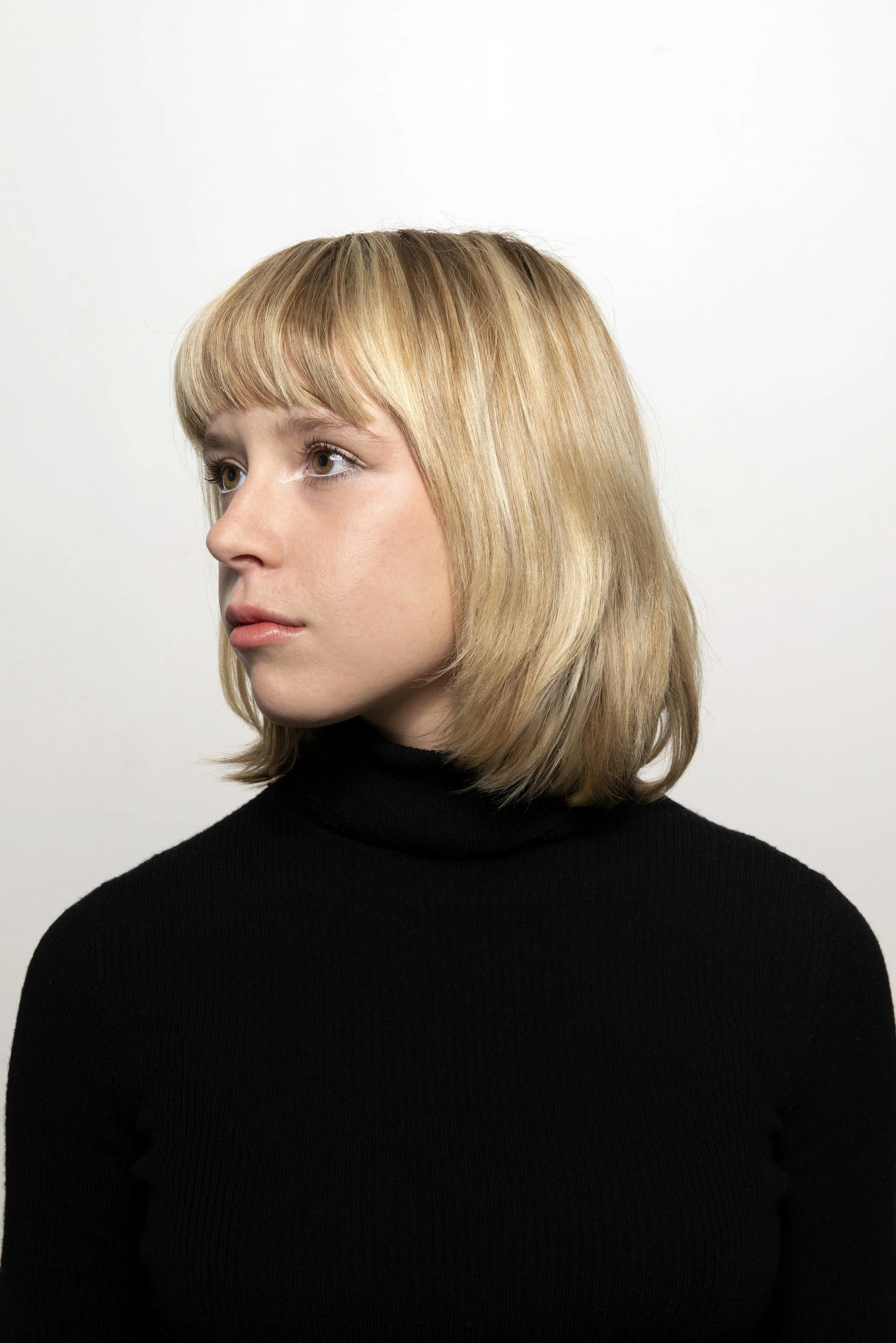 A woman with blonde hair and light skin, wearing a black turtleneck, looking to the side against a plain white background.