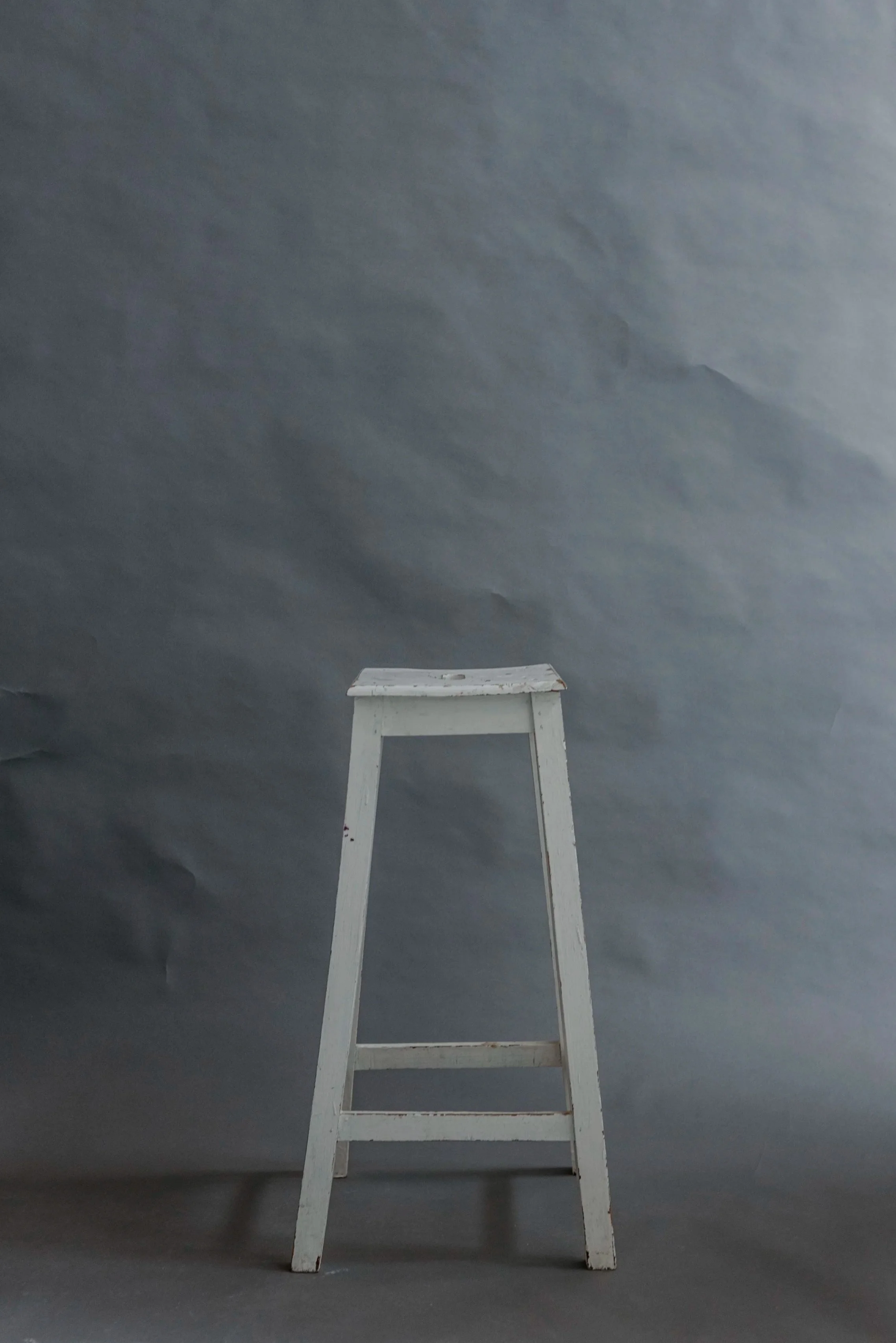 A white wooden stool with a worn top sits on a gray floor against a dark gray, textured background.