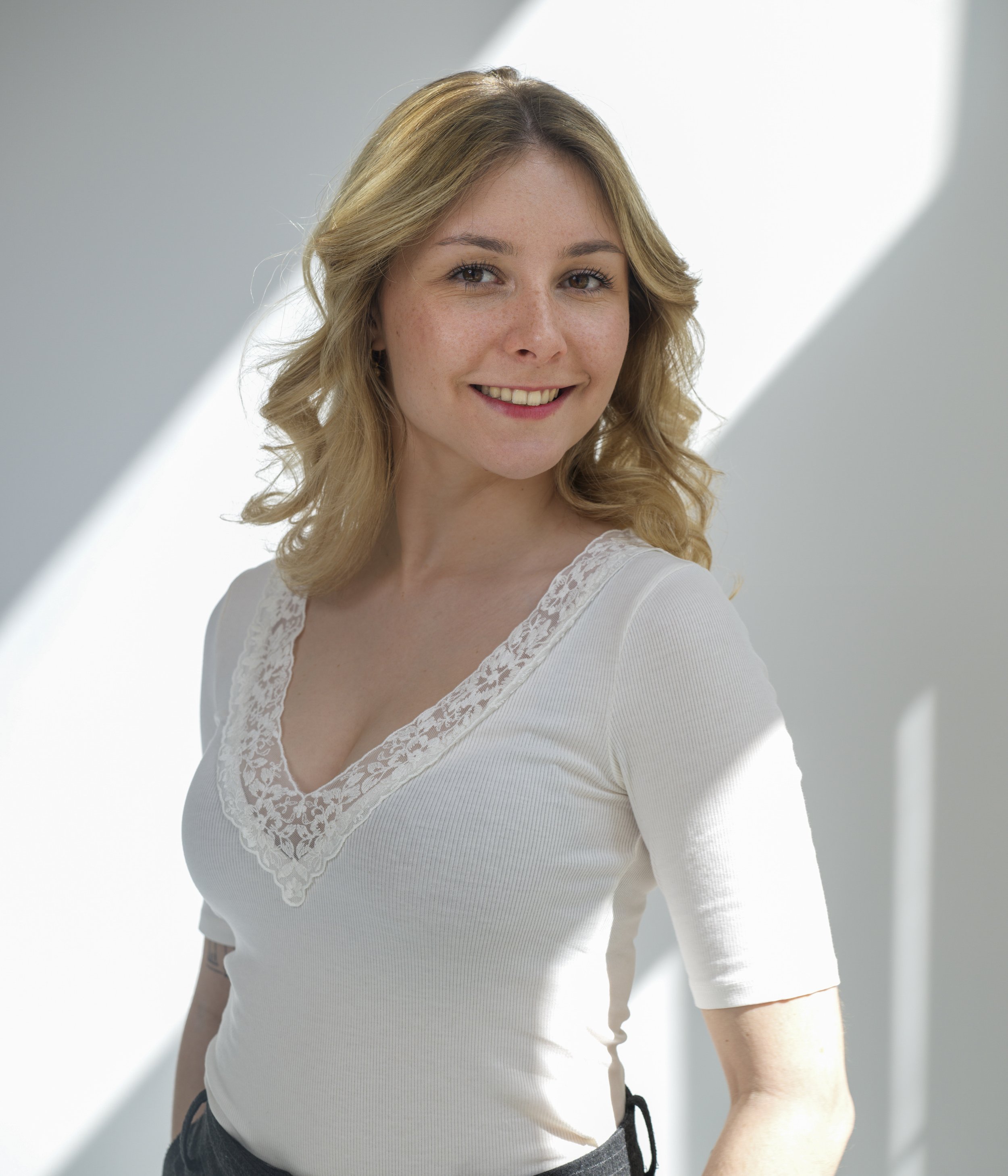 A young woman with blonde wavy hair and light skin smiling outdoors, wearing a white top with lace details around the neckline.