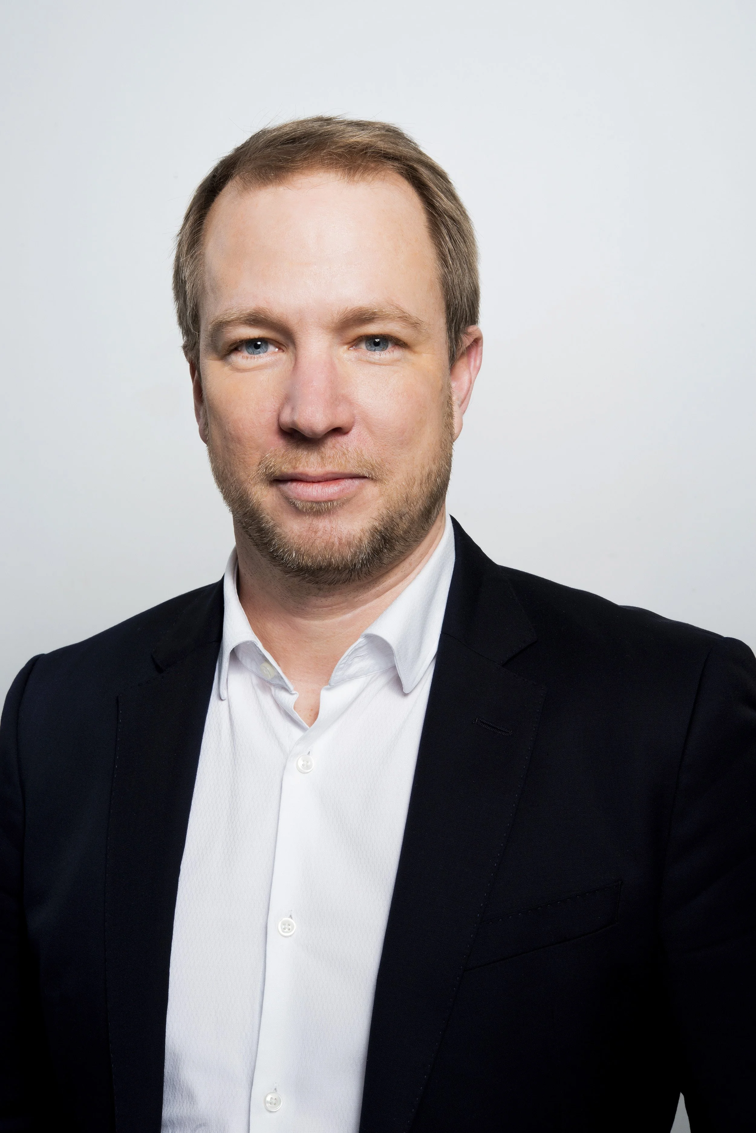 Headshot of a man with light brown hair and blue eyes, wearing a black blazer and white shirt, against a plain, light-colored background.