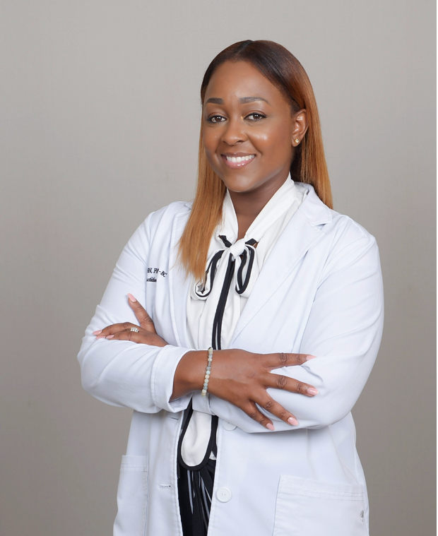 A confident woman wearing a white lab coat with a black and white scarf, smiling, with arms crossed, standing in front of a plain gray background.