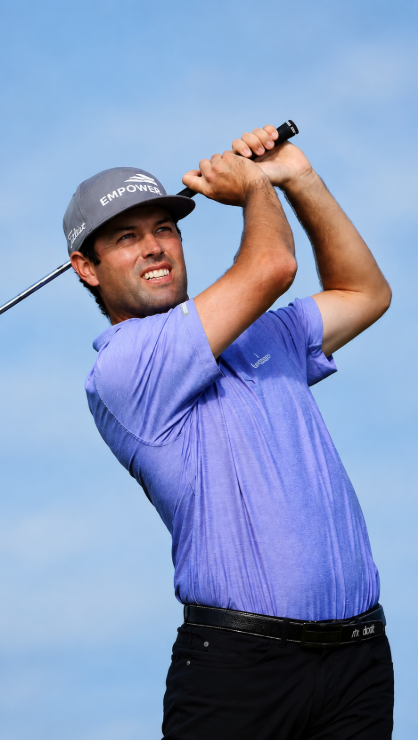 Male golfer wearing a gray hat and blue polo shirt, mid-swing against a blue sky background.