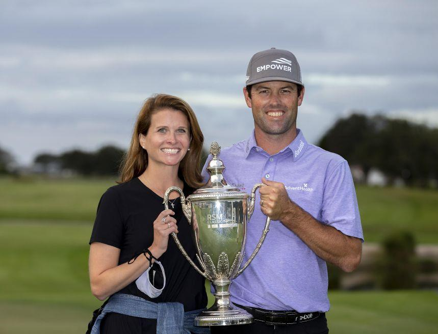 A man and woman holding a large silver trophy together on a golf course, both smiling, with trees and cloudy sky in the background.