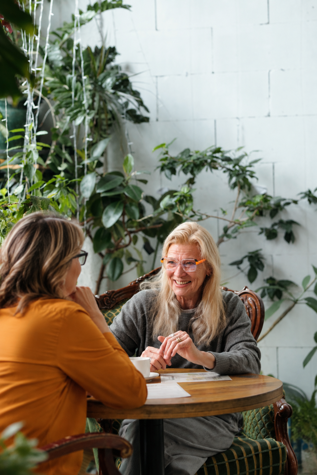 Two women sitting at a wooden table, engaging in conversation, with lush green plants and a white brick wall in the background.