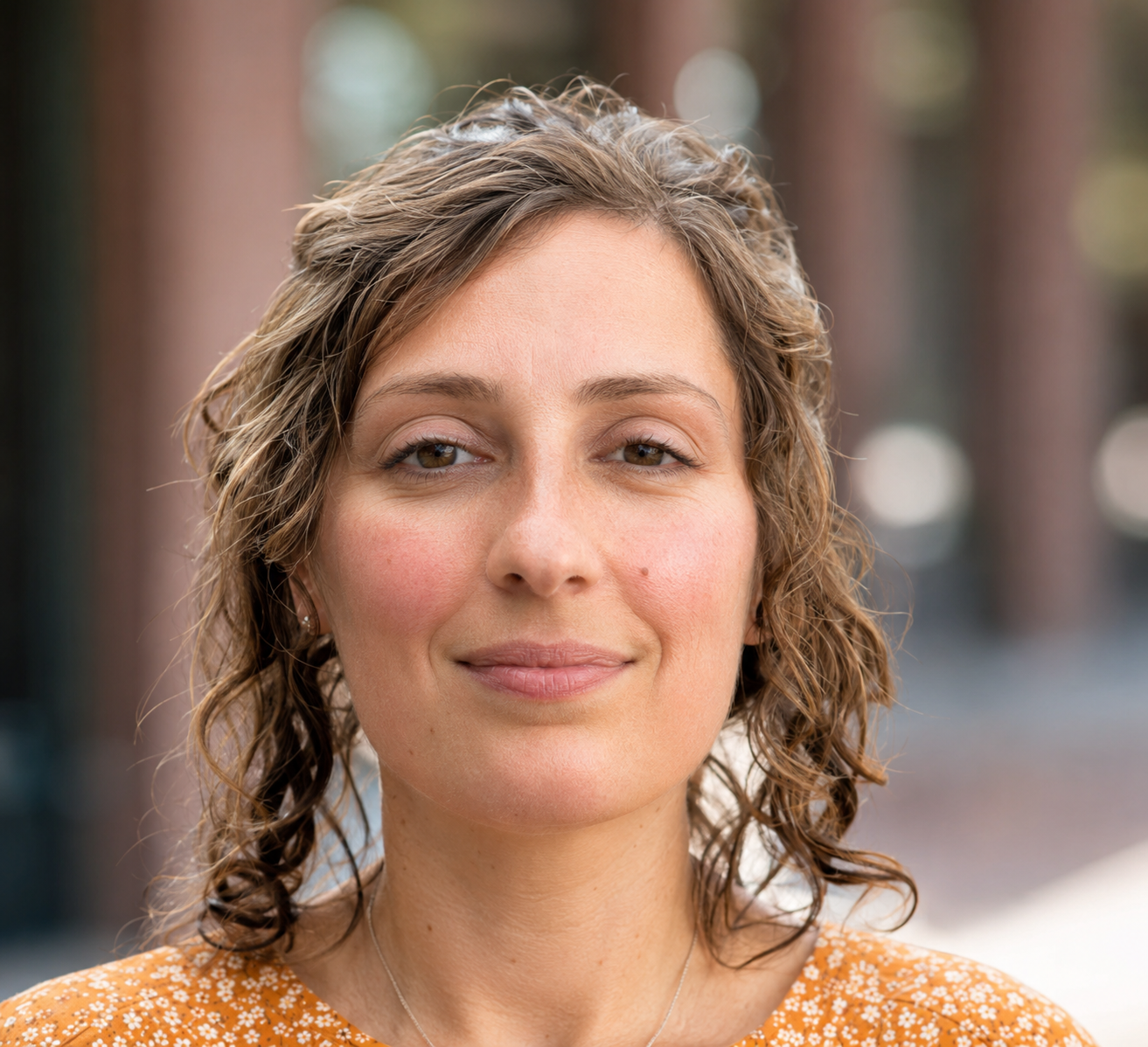 A close-up of a smiling woman with curly brown hair, wearing a yellow floral blouse, standing outdoors with blurred trees in the background.