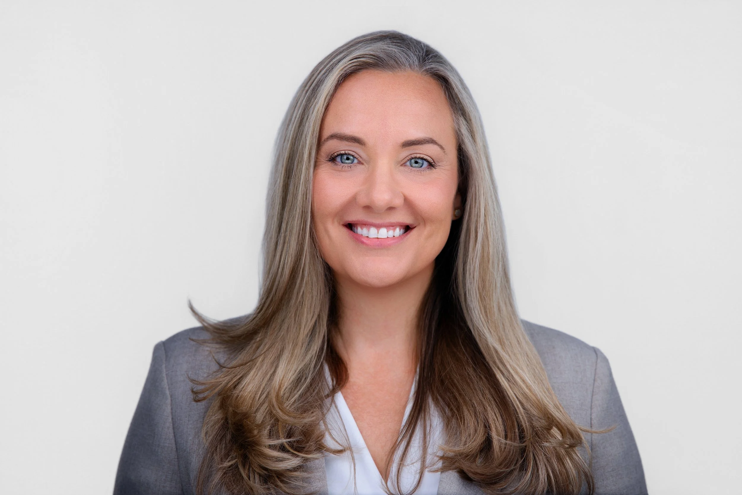 A woman with long, wavy blonde hair, blue eyes, and a bright smile, dressed in gray business attire, standing against a plain white background.