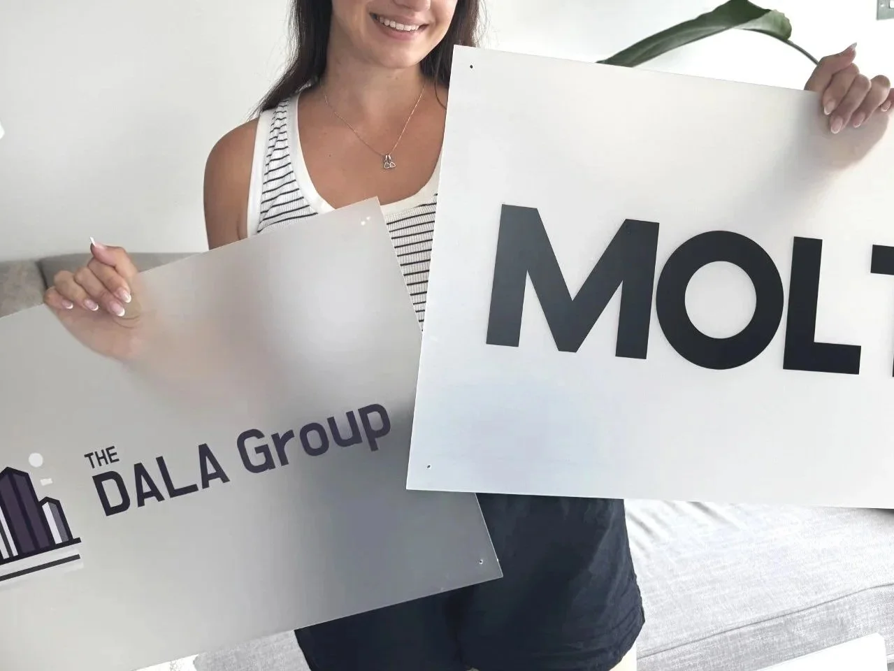 A smiling woman holding two signs, one says "THE DALA Group" and the other says "MOL".
