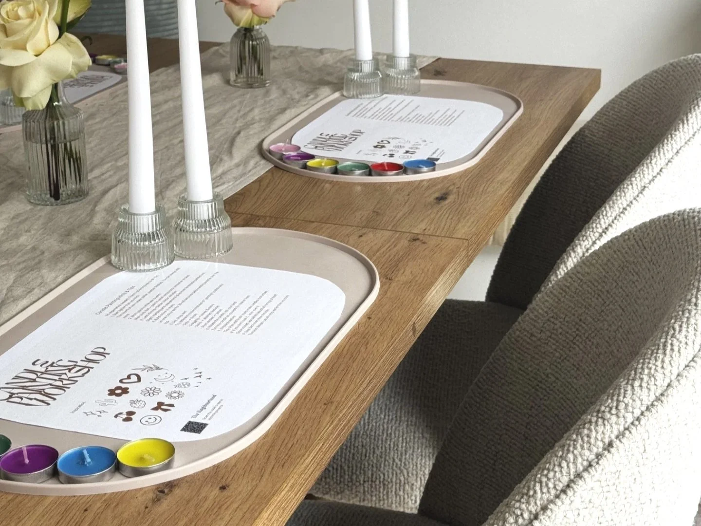 A dining table set with two beige trays holding paper placemats, tea candles, and a paint palette with watercolors. Two glass vases with flowers and three white candlesticks are also on the table, with upholstered chairs surrounding it.
