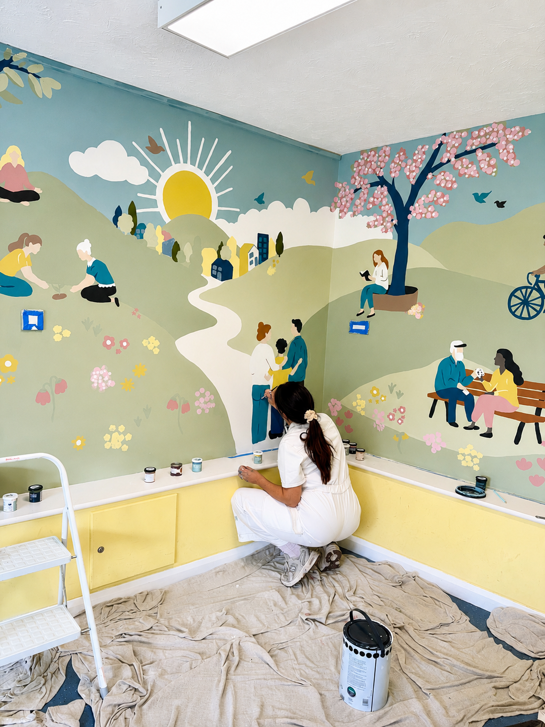 A woman in white overalls painting a colorful mural on a corner wall, depicting a sunny landscape with people, animals, trees, and flowers.