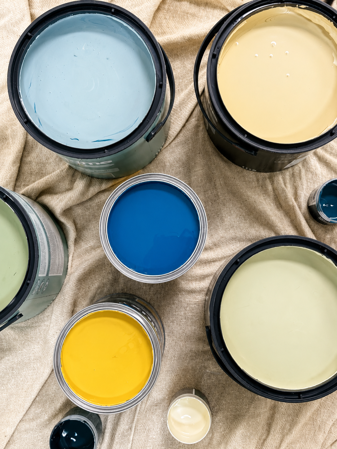 Multiple buckets of paint in various colors including blue, yellow, green, and cream, displayed on a beige fabric surface.