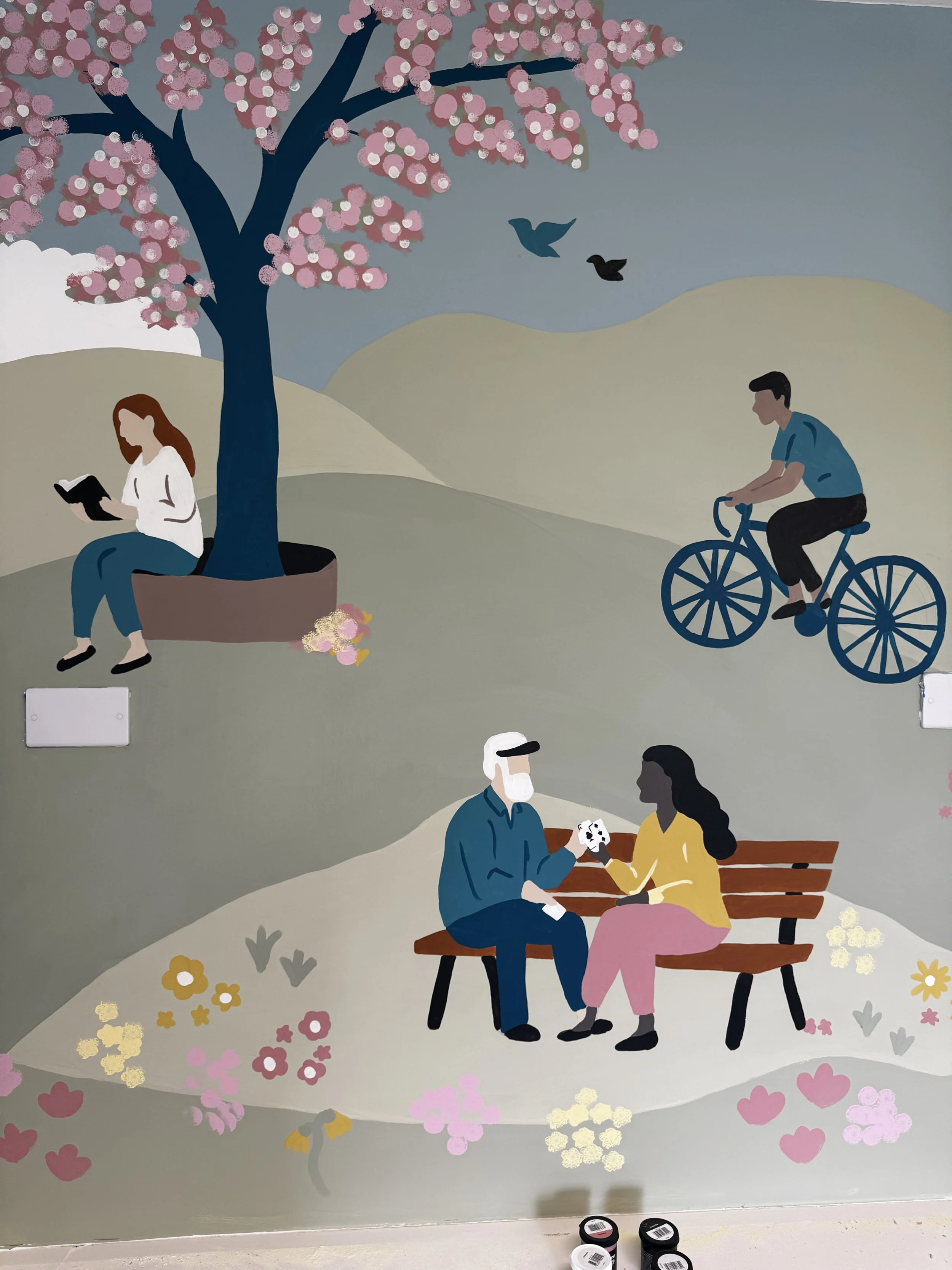 A mural depicting a park scene with trees, blossoming flowers, and various people doing different activities: a woman sitting under a tree reading a book, a man riding a bicycle, and a couple sitting on a bench talking. Two birds fly in the sky, and 