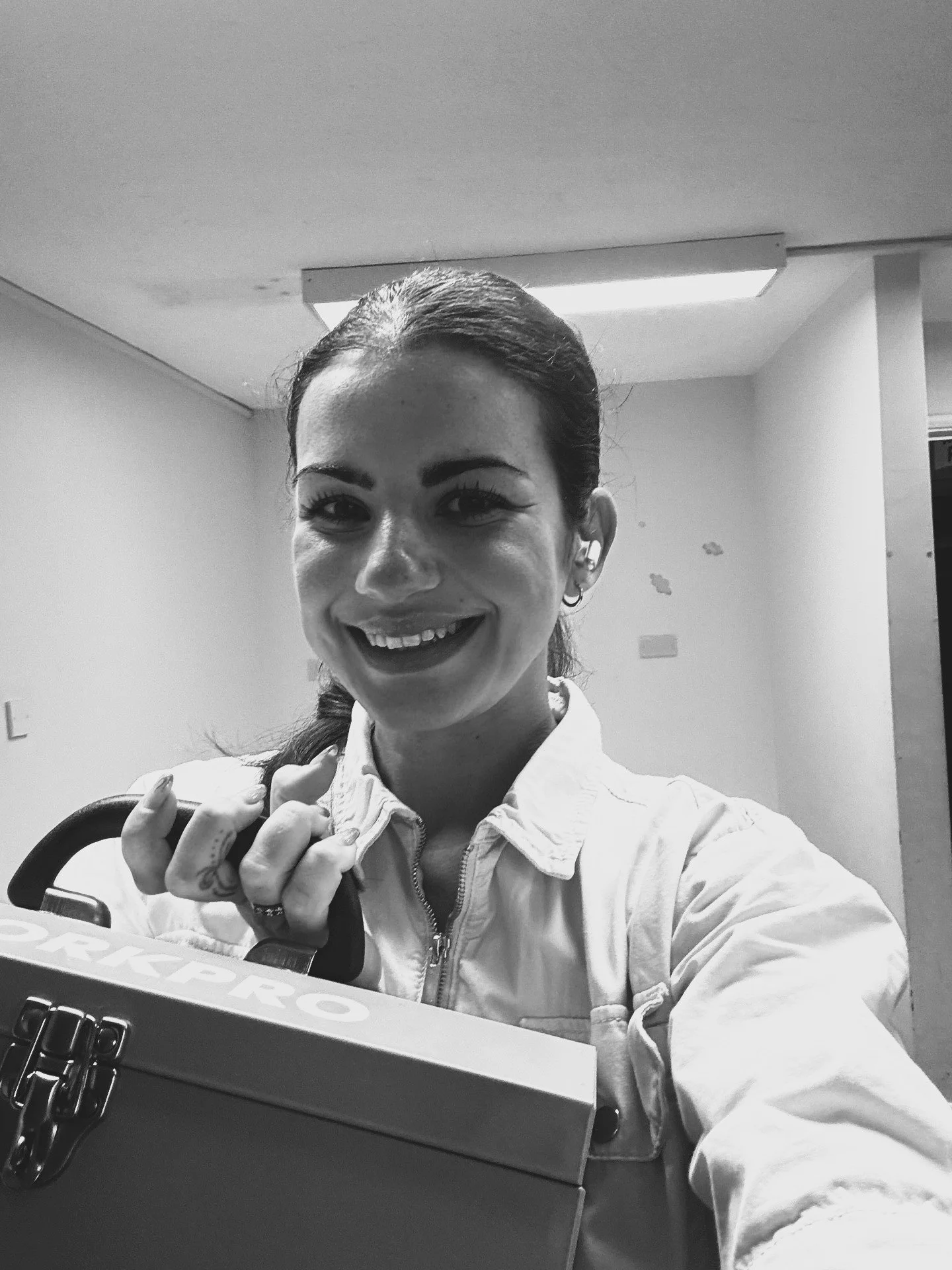 A smiling woman with dark hair tied back, holding a toolbox full of artistic paintbrushes, standing in a room that she is about to paint, there is a fluorescent light on the ceiling.