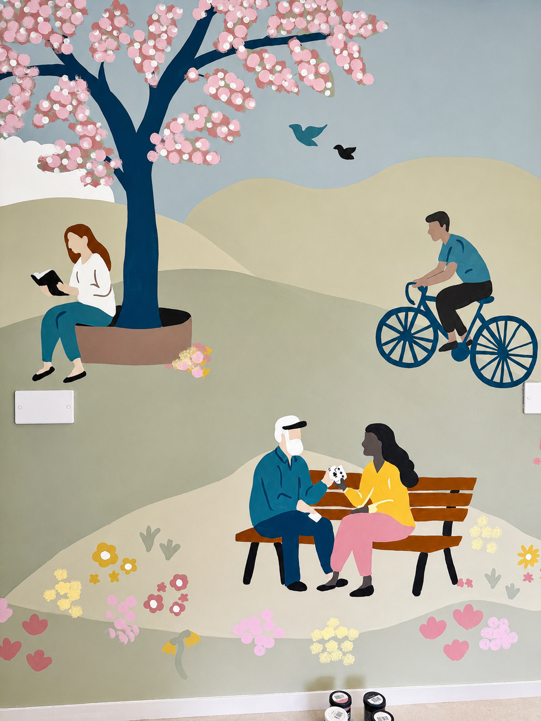 A mural painting depicting a park scene with people: a woman reading under a cherry blossom tree, a man riding a bicycle, and an elderly man and woman sitting on a bench with a dog.