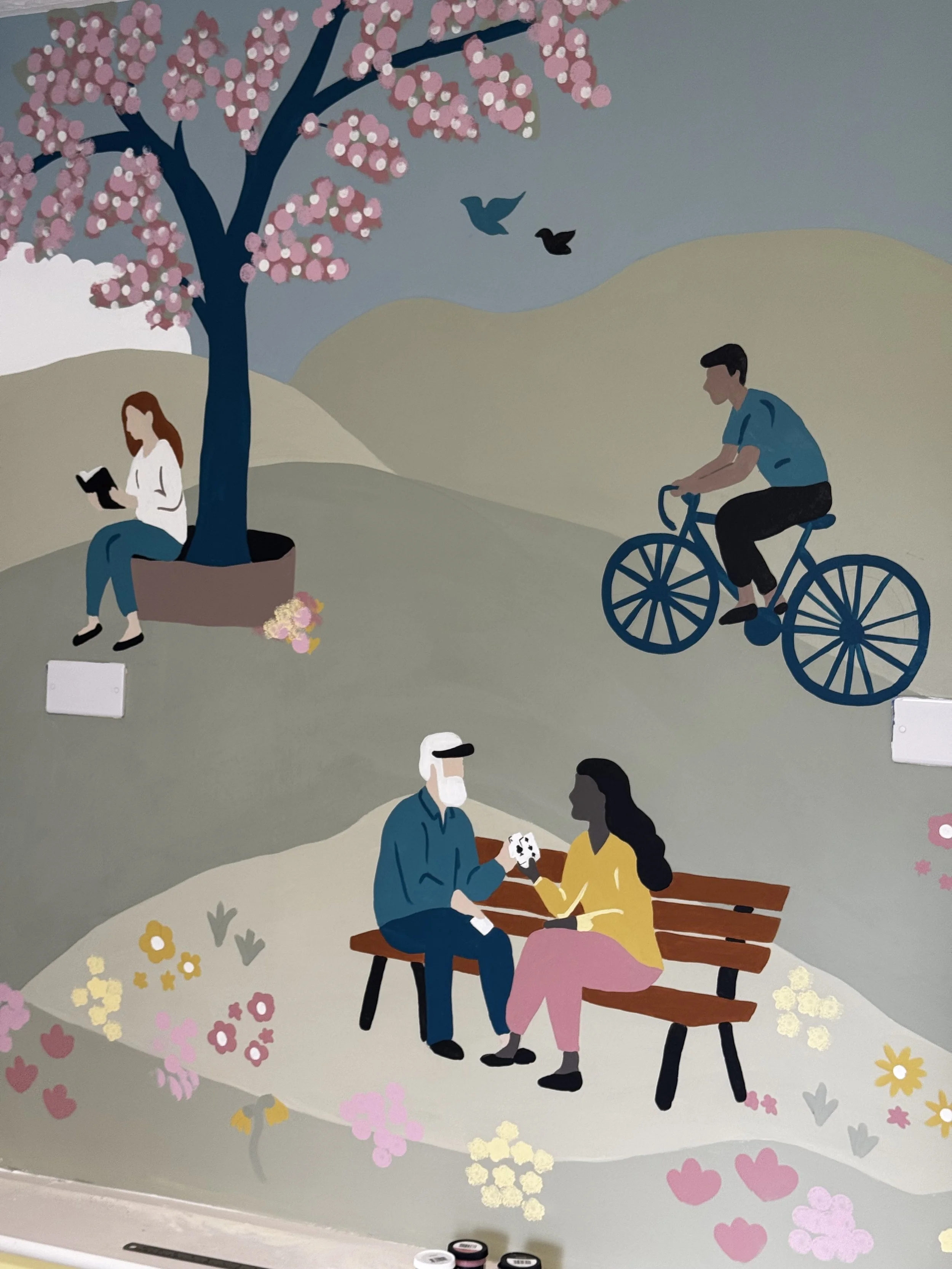 A stylized mural featuring a pink blossoming cherry tree, a woman sitting on a bench reading, a man and woman sharing a kiss at a picnic, a cyclist riding past, and birds flying in the sky. Flowers and hills are in the background.
