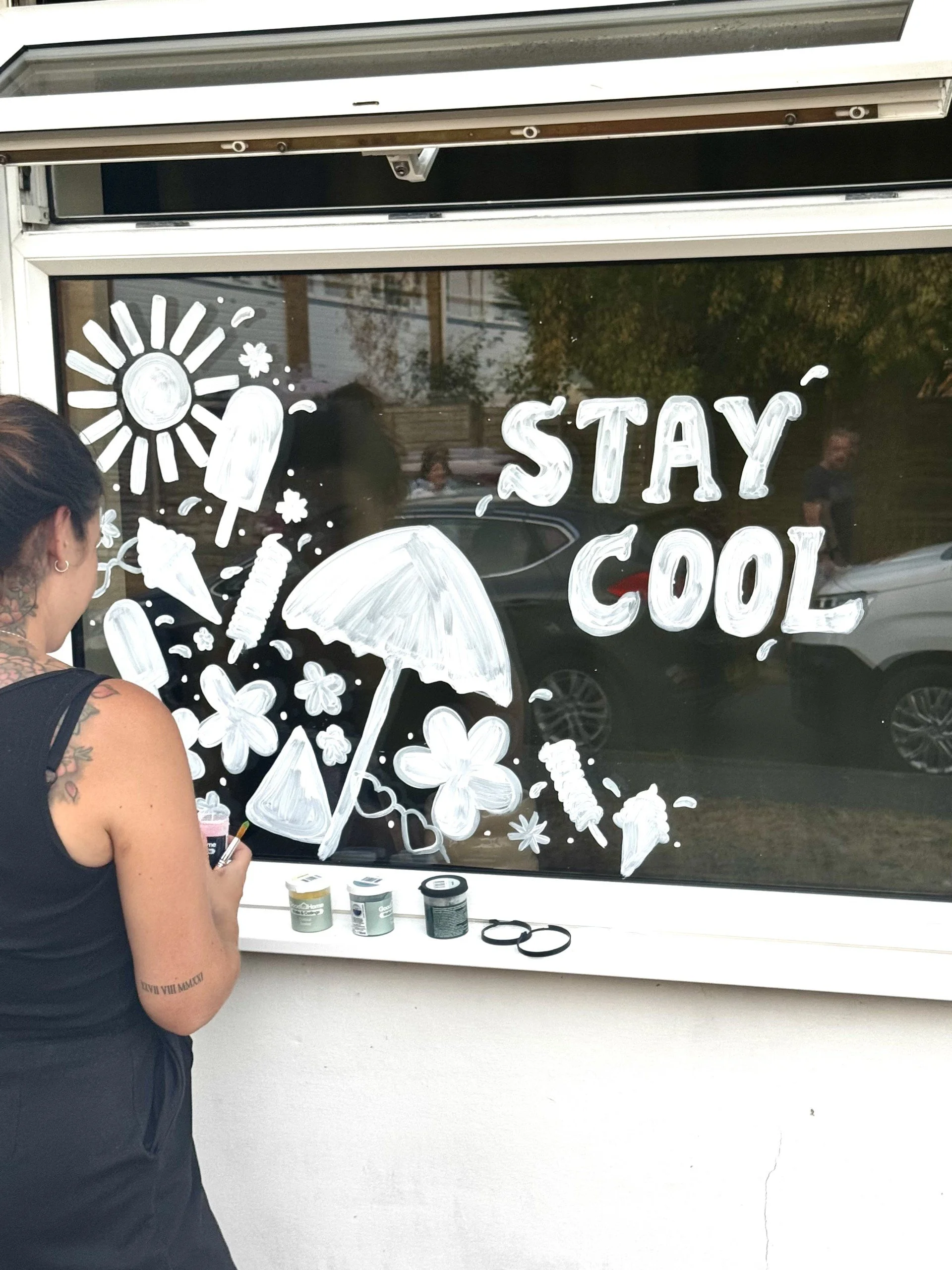 A person painting white illustrations and the words 'STAY COOL' on a window, including drawings of ice cream, popsicles, umbrellas, and flowers.