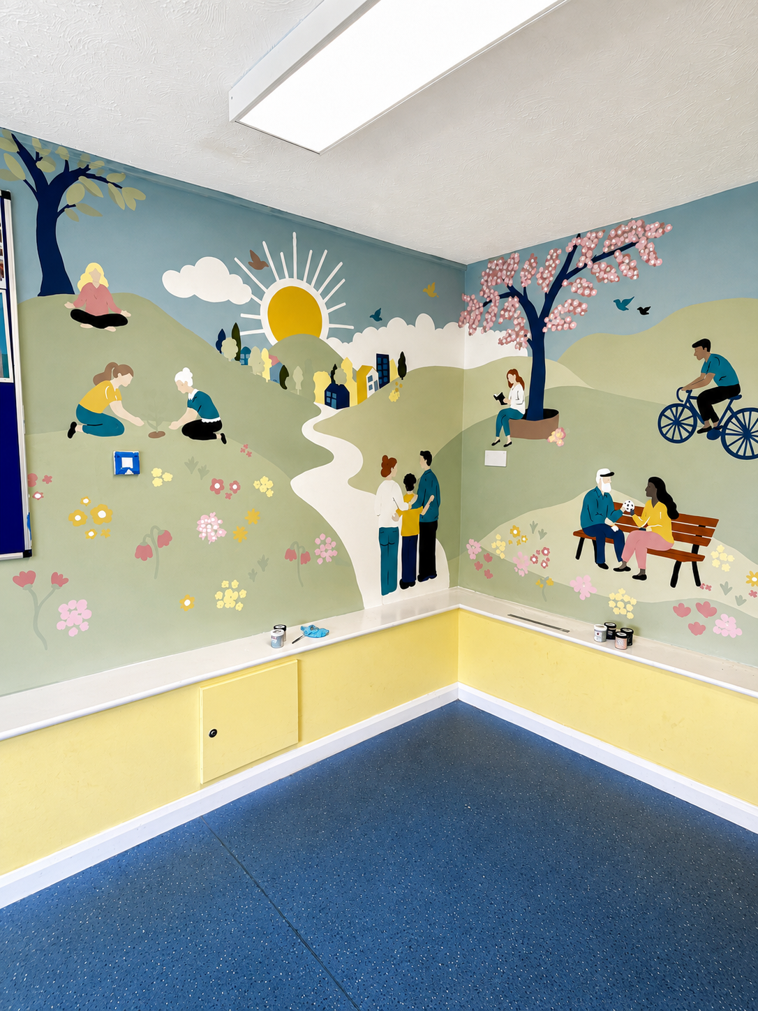 Colorful indoor wall mural depicting a park scene with children and adults engaging in activities like playing, reading, riding bikes, and talking. The mural features a sunny sky, rolling green hills, trees with pink blossoms, flowers, and a pathway through the park.