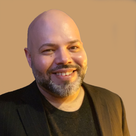 A smiling bald man with a beard, wearing a black shirt and a dark blazer, against a plain beige background.