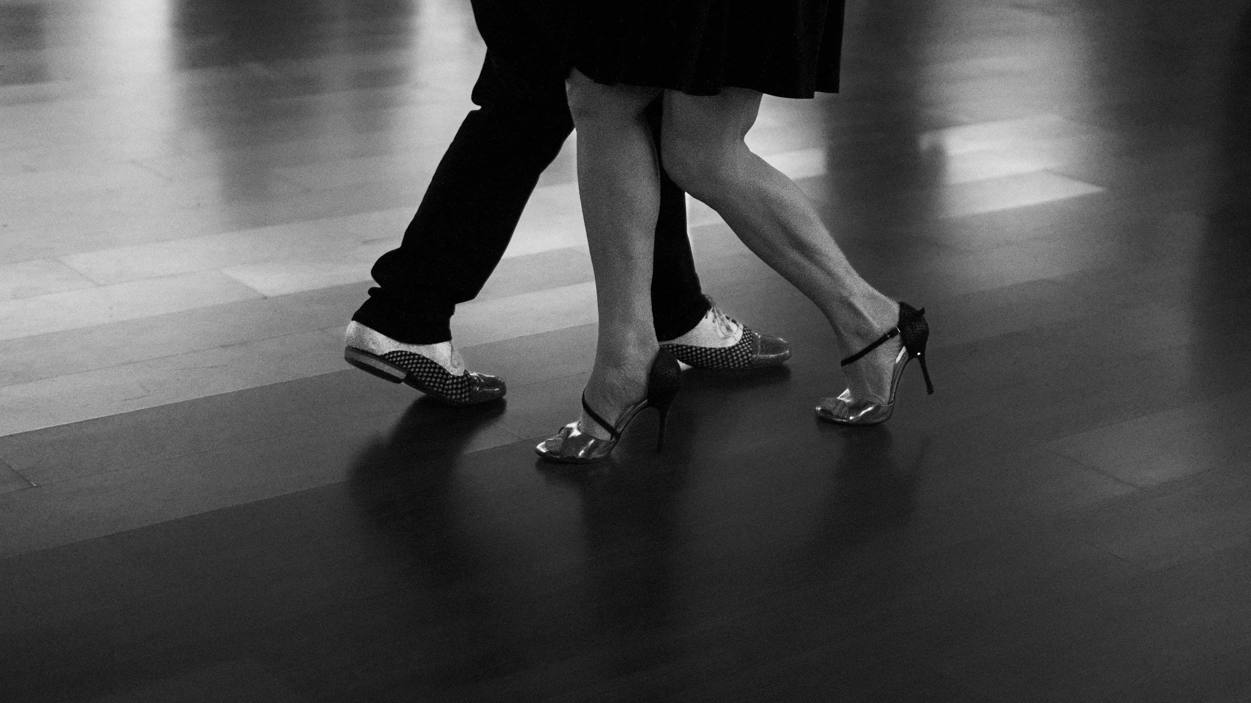 A black and white image of a couple dancing; the woman is wearing high heels and a skirt, and the man is wearing dress shoes and dark pants.