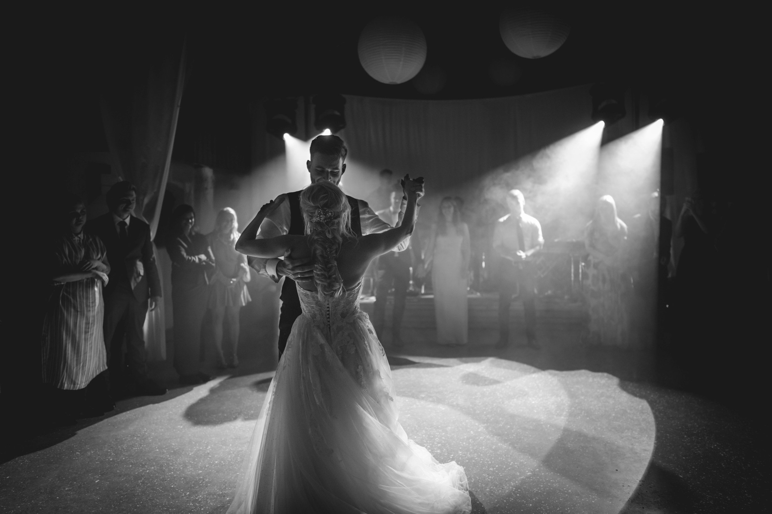 A black and white photo of a couple dancing closely at a wedding reception, surrounded by guests and soft lighting.