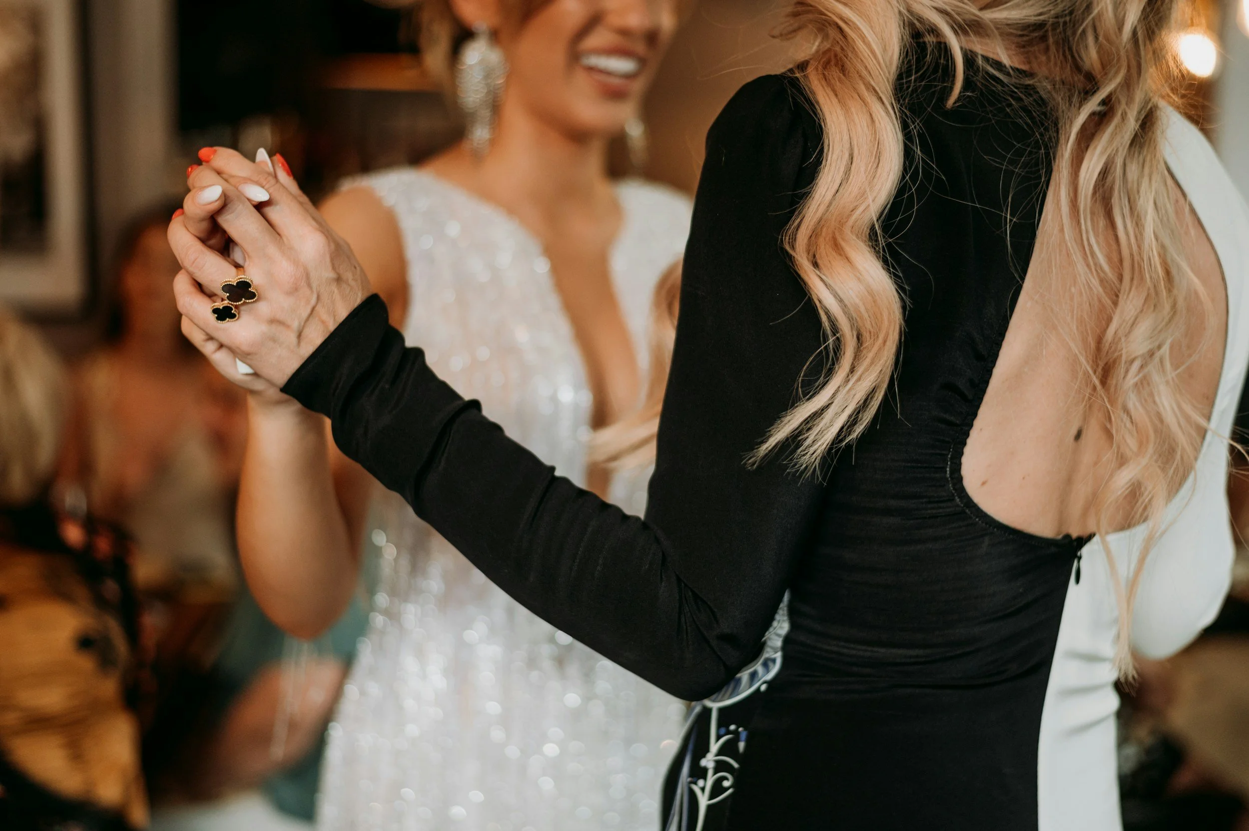 Two women holding hands and dancing at a celebration or wedding, with one woman wearing a white dress and the other wearing a black top.