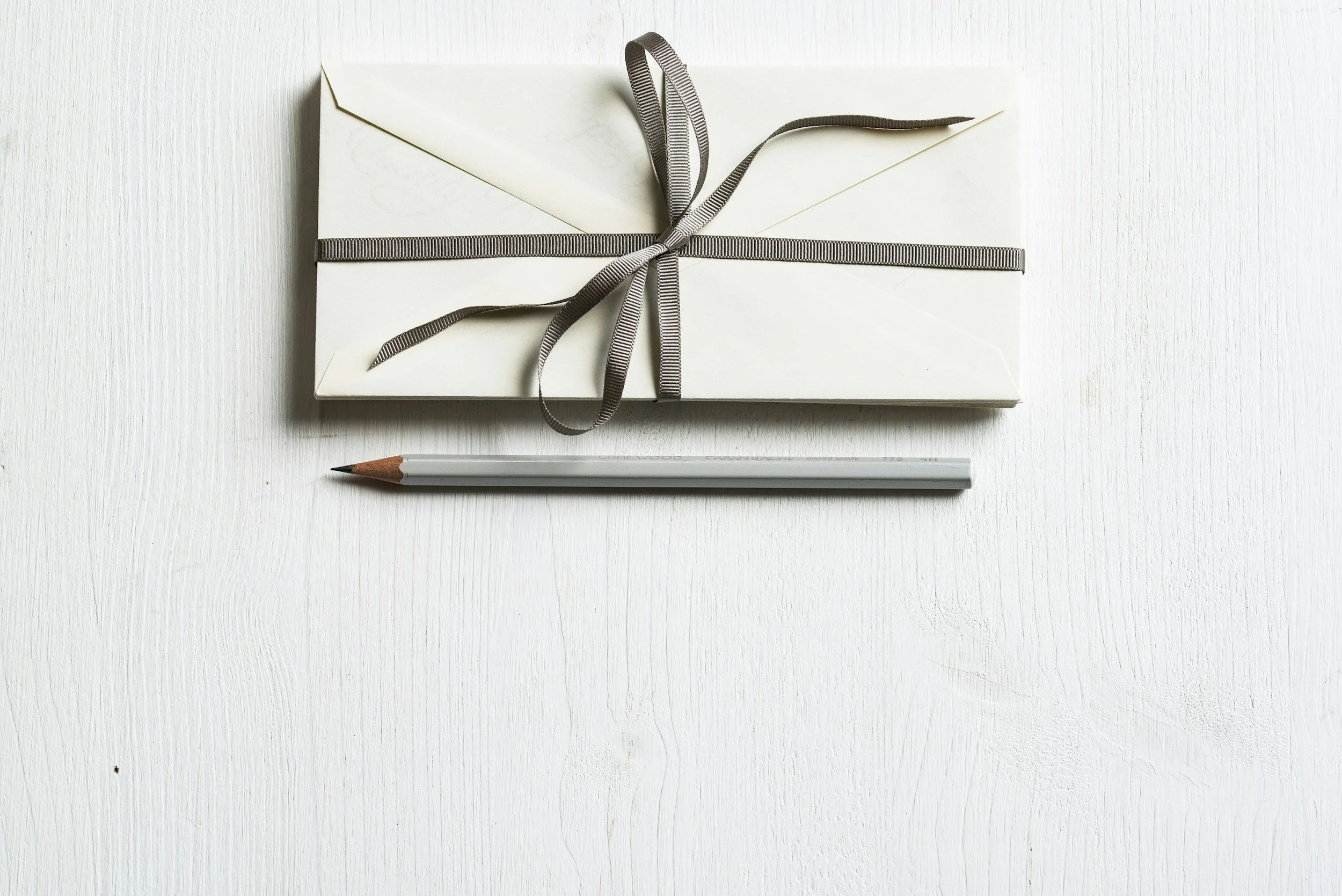 A white envelope tied with a gray ribbon, placed on a white wooden surface with a gray pencil underneath.