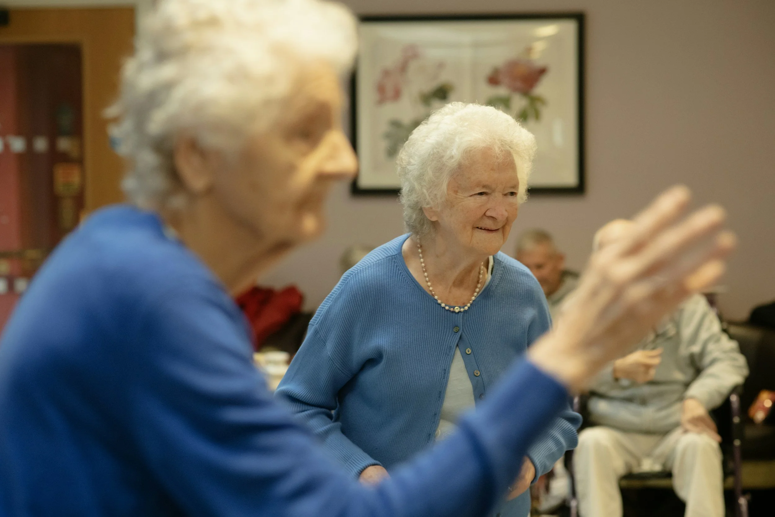 An elderly woman with white hair and a pearl necklace, smiling and sitting in a room with other people, during a social gathering or event.