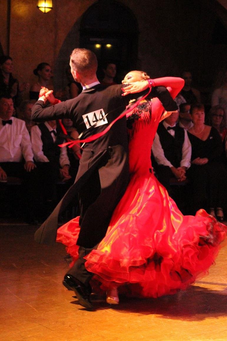 A couple dancing, with the woman wearing a red gown and the man in a black suit with the number 144 on his back, during a ballroom dance competition.