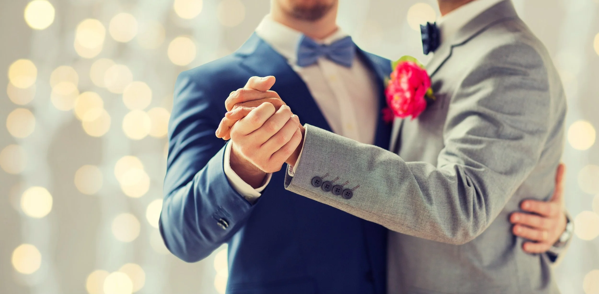 A couple dancing at a wedding reception, holding hands and embracing each other. The focus is on their hands and upper bodies, with the background blurred with warm bokeh lights.