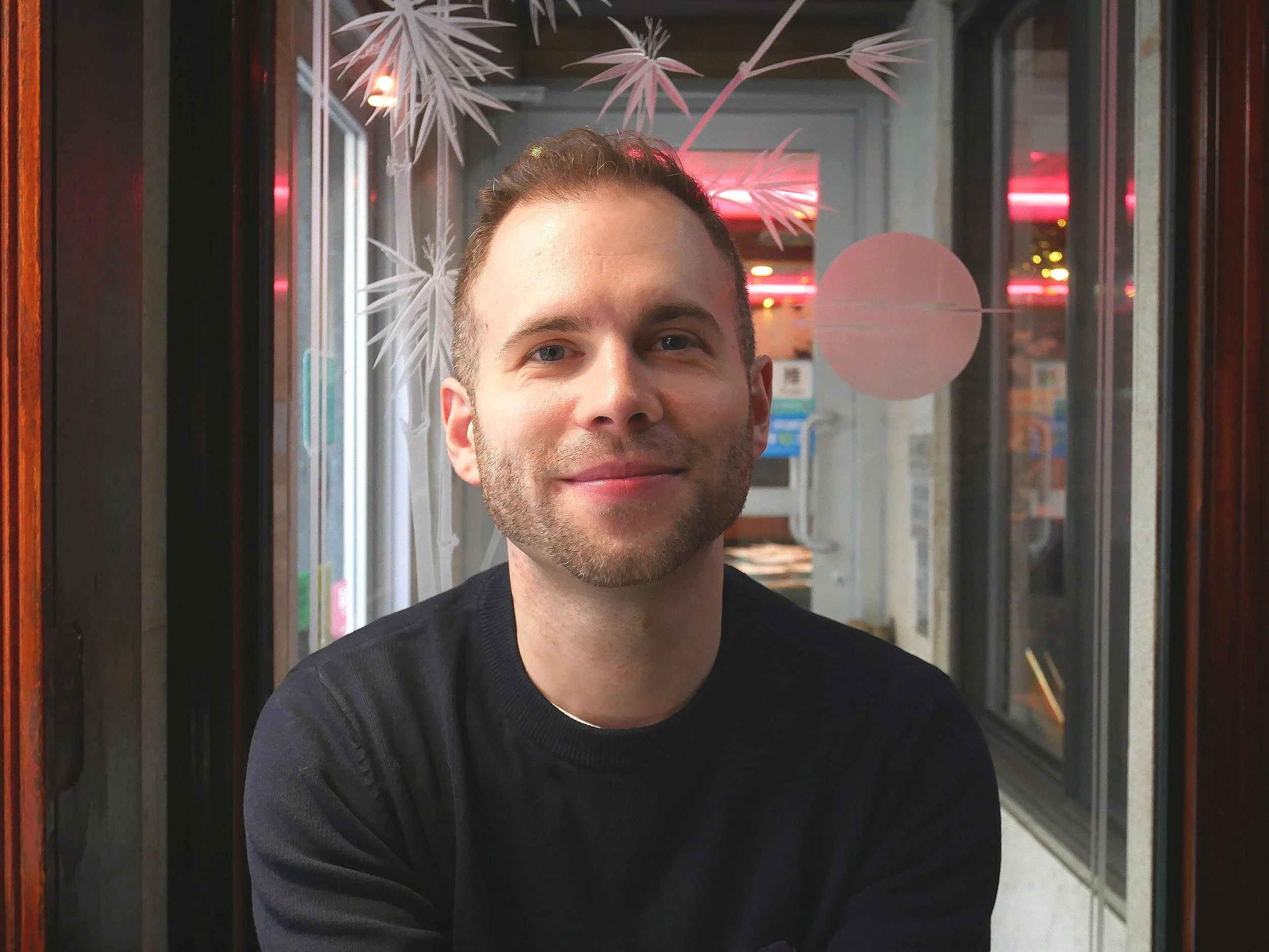 A man with short, light brown hair and a slight beard smiling at the camera. He is wearing a black shirt and is standing in front of a window with Christmas-themed decorations, including paper snowflakes and a pink circle.