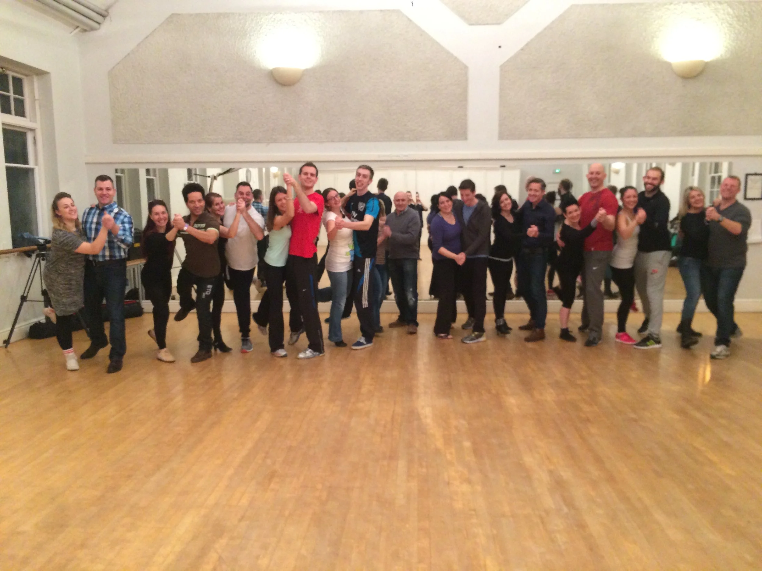 Group of people dancing and smiling in a dance studio with a wooden floor and a large mirror on the wall.