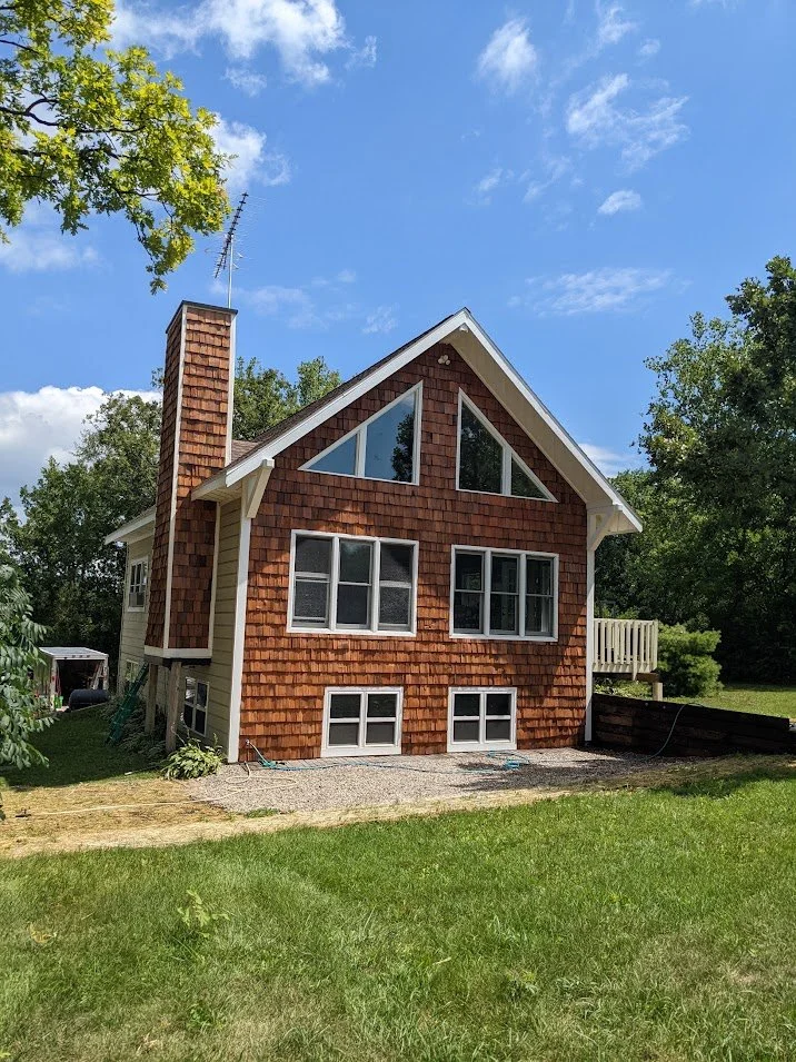 A two-story house with a steep pitched roof, shingle siding, multiple windows, and a small wooden deck, surrounded by green trees and grass under a blue sky.