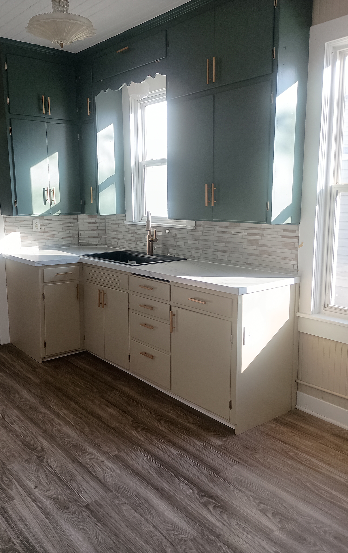 A kitchen with dark green upper cabinets, white lower cabinets, a white countertop, a black sink, a window above the sink, a light fixture on the ceiling, and wood-patterned flooring.