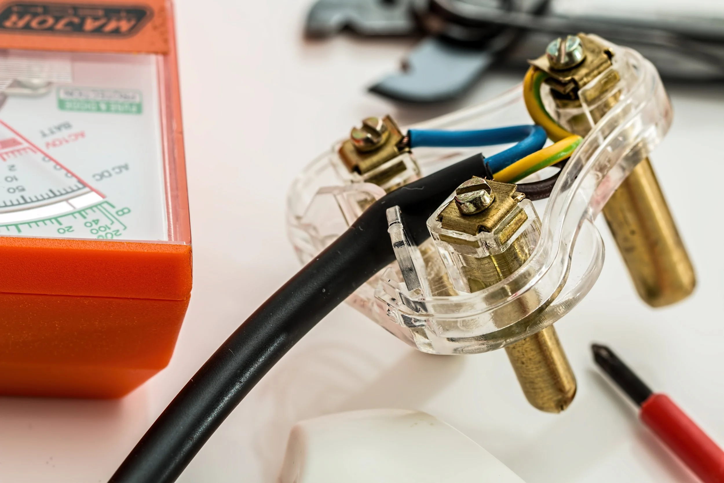 Close-up view of electrical wiring with black, blue, and yellow wires connecting to a transparent socket, with an orange toolbox or component partially visible on the left side and a white surface underneath.