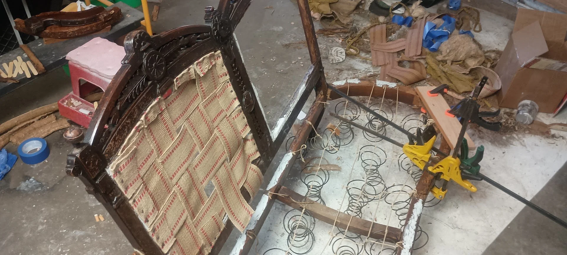 Photo of a partially disassembled vintage wooden bed frame with decorative headboard, with coil springs and tools, in a cluttered workshop with cardboard and packaging materials on the floor.