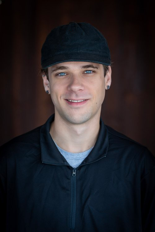 A young man wearing a black cap, black jacket, and gray shirt, smiling at the camera against a dark background.