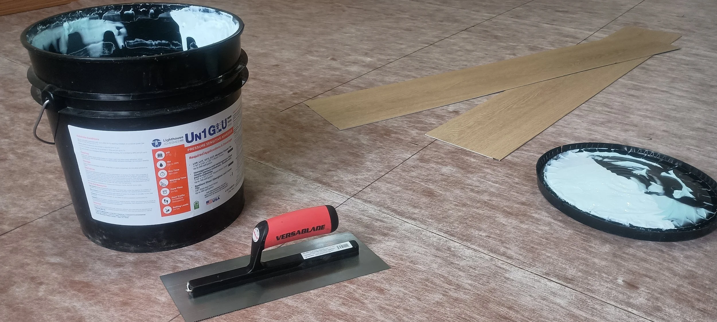 Tools for installing flooring on a wooden surface, including a bucket of adhesive, a notched trowel, and a roll of flooring planks.