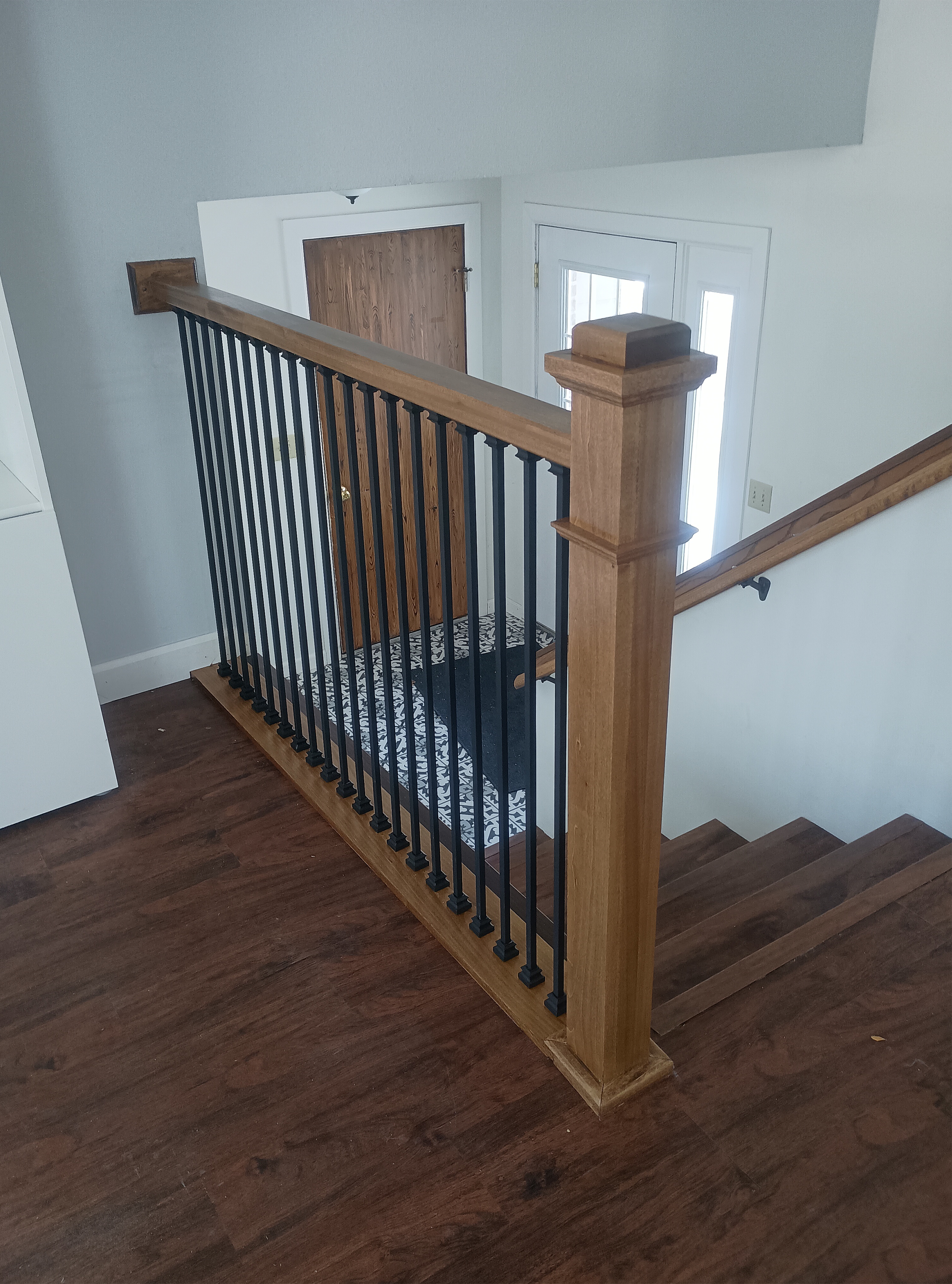 Interior view of a staircase with a wooden handrail, black metal balusters, and a patterned carpet runner, leading to a doorway with a wooden door and a small window in a house.