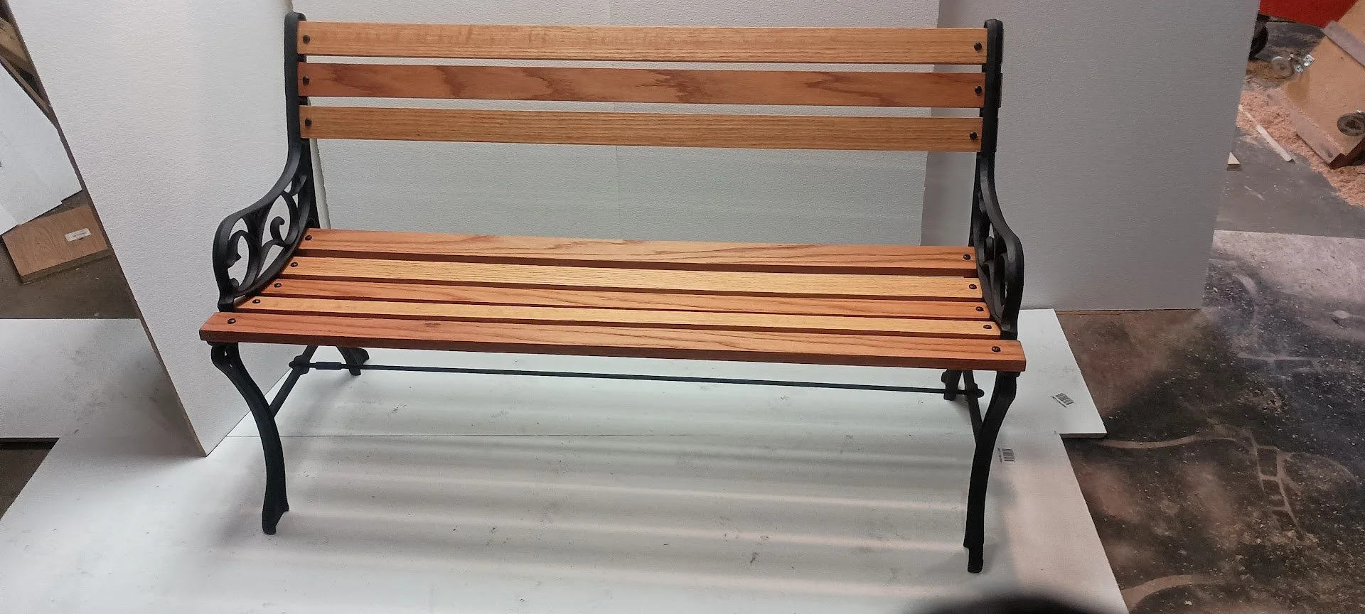 Wooden slat outdoor bench with black metal armrests and legs, situated on a white surface in a store or warehouse setting.