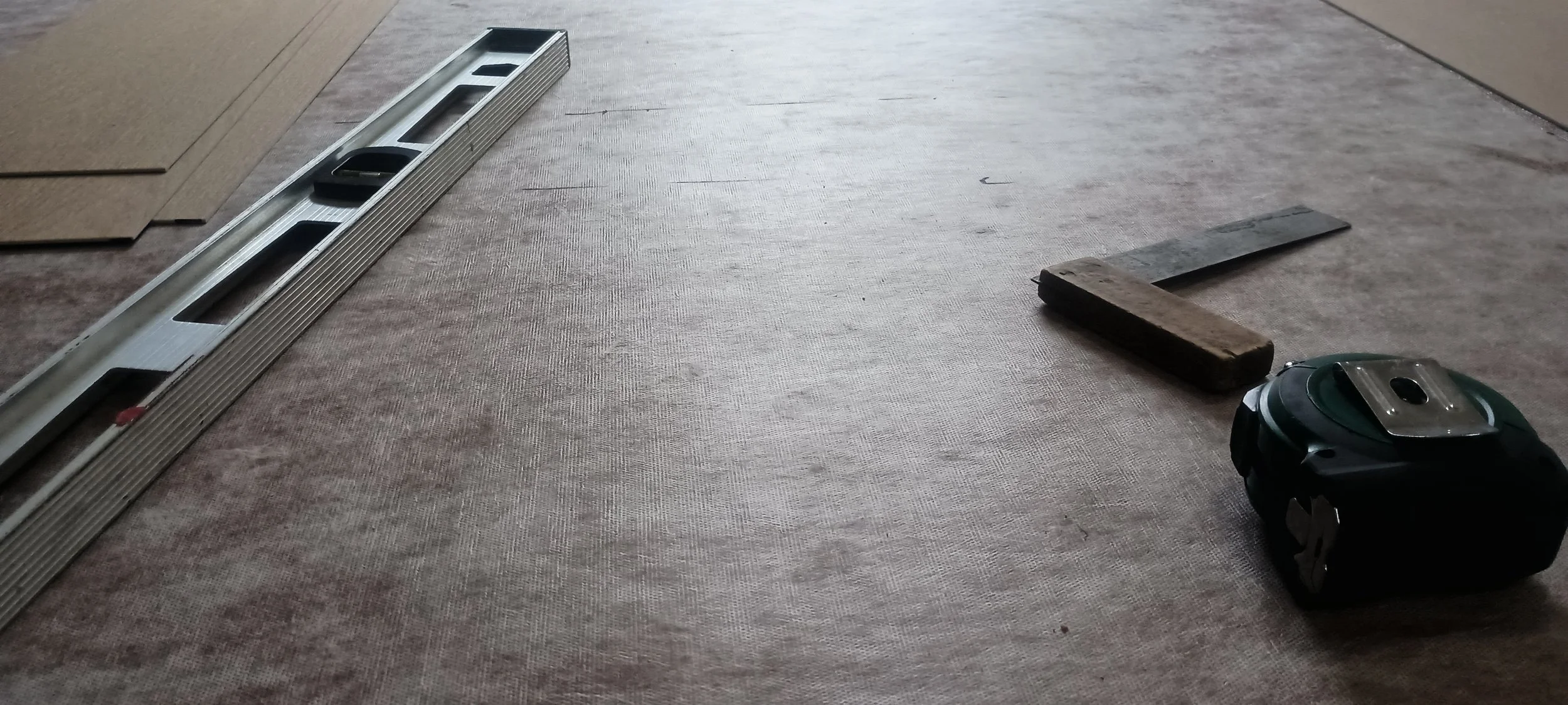Construction tools and materials on a floor, including a spirit level, a wooden plank, a metal ruler, and a power tool.