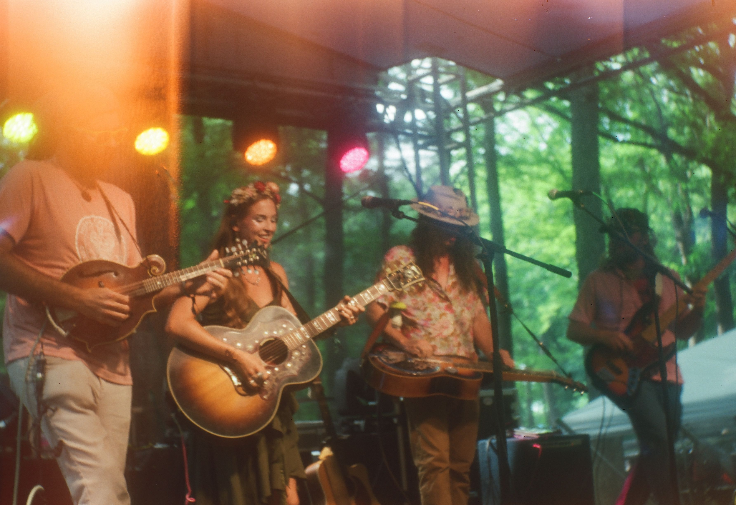 Group of musicians playing guitars and singing on a stage with a forest background, colorful stage lights illuminating the scene.