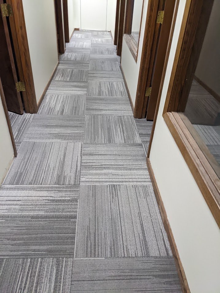 An indoor corridor with gray patterned carpet tiles on the floor, white walls, and wooden doorframes and window sills.