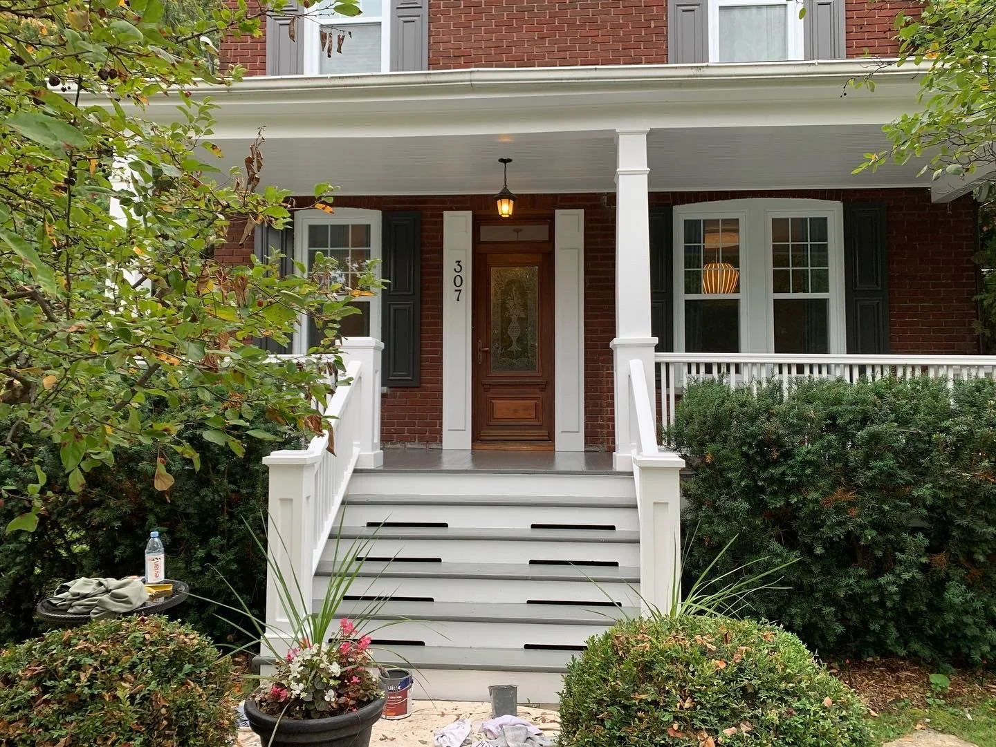 Front porch of a brick house with white railings and stairs, a wooden door, and black window shutters. The house number is 307. There are shrubs, plants, and a large potted plant in front.