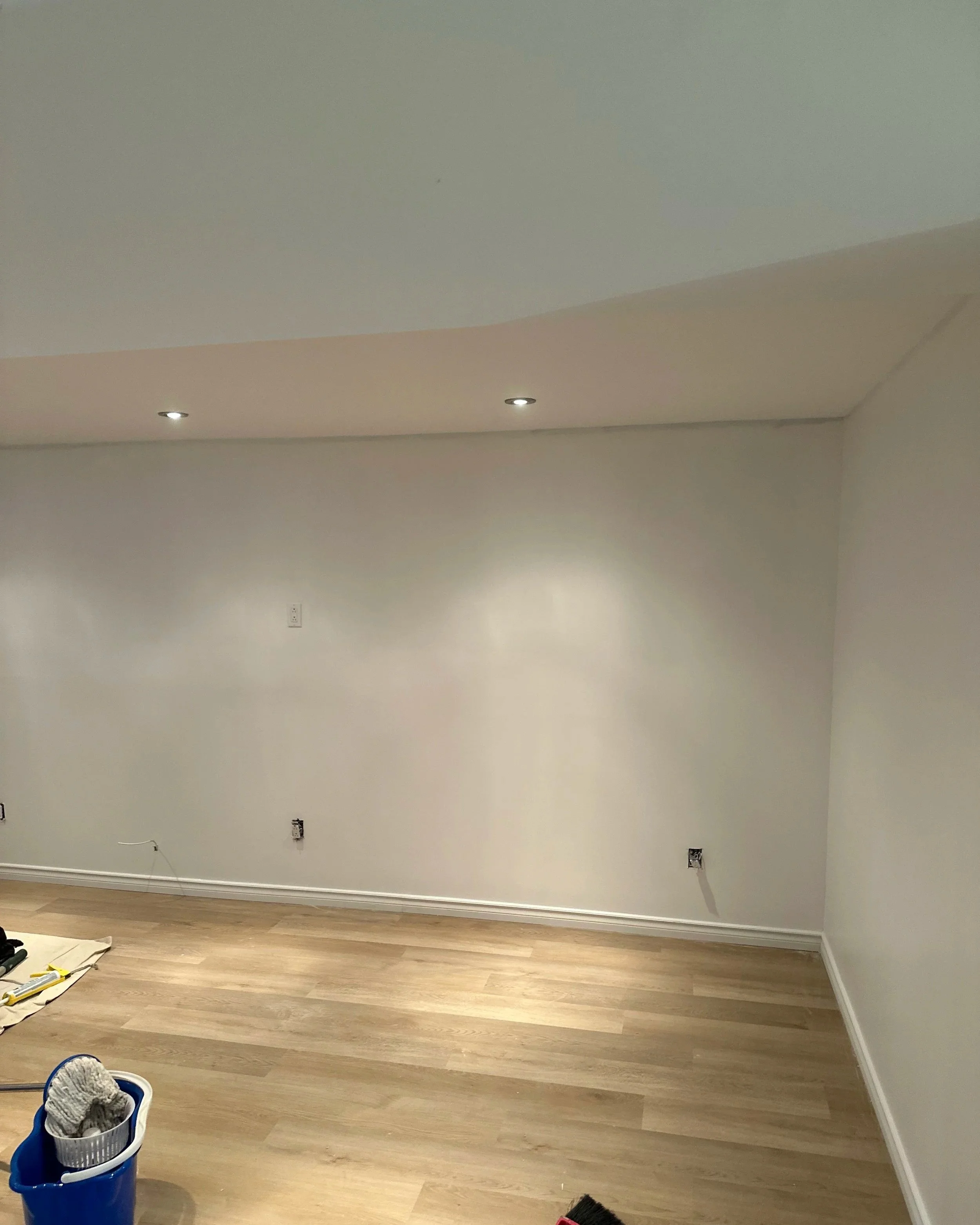 A partially renovated room with beige wooden flooring and white walls. Several electrical outlets are visible on the wall. Cleaning tools, including a broom with a red handle and a blue bucket with cleaning supplies, are on the floor. Recessed lighti
