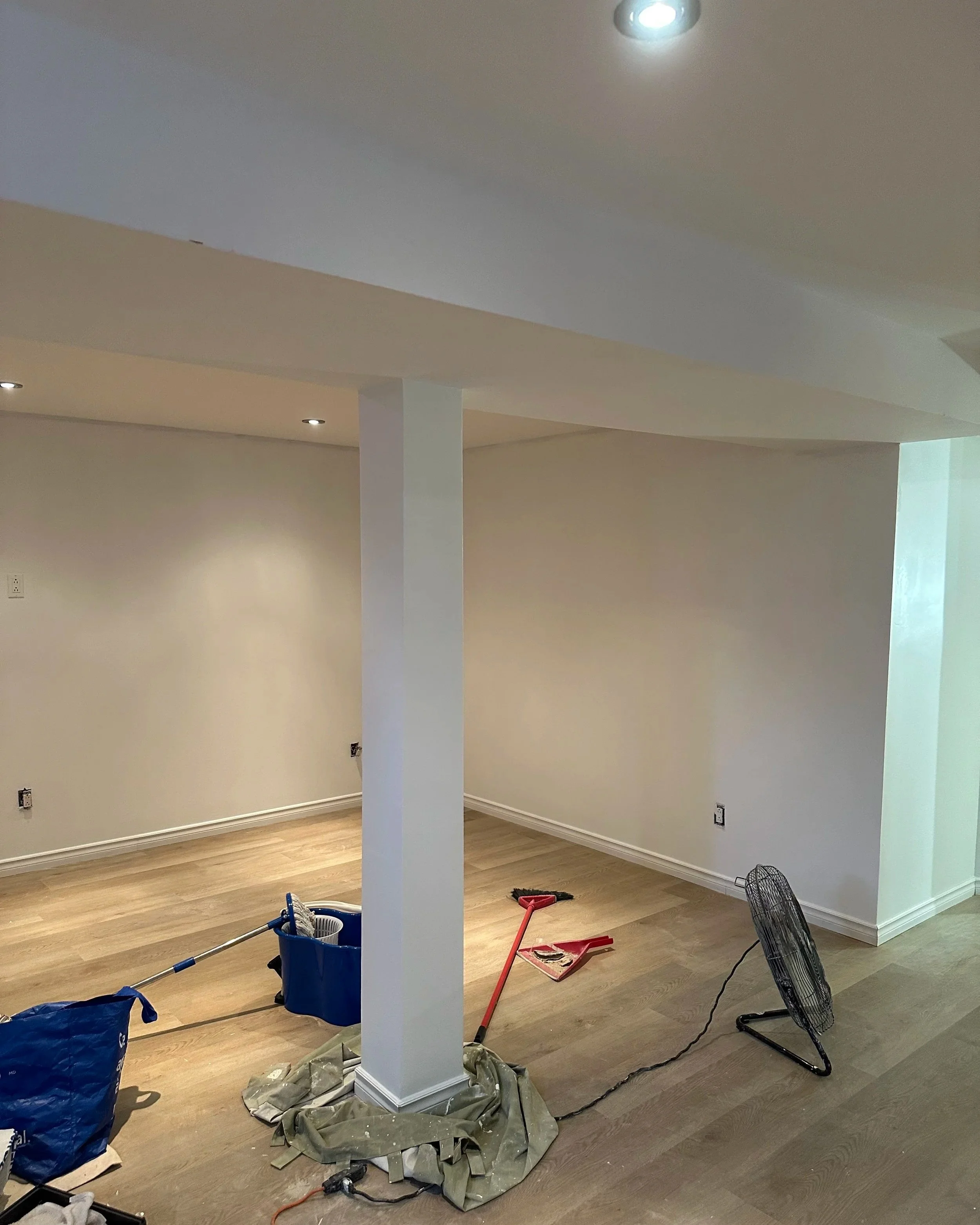 Interior room under renovation with tools on the floor, including a red trowel, black fan, and blue bucket, and scattered materials.
