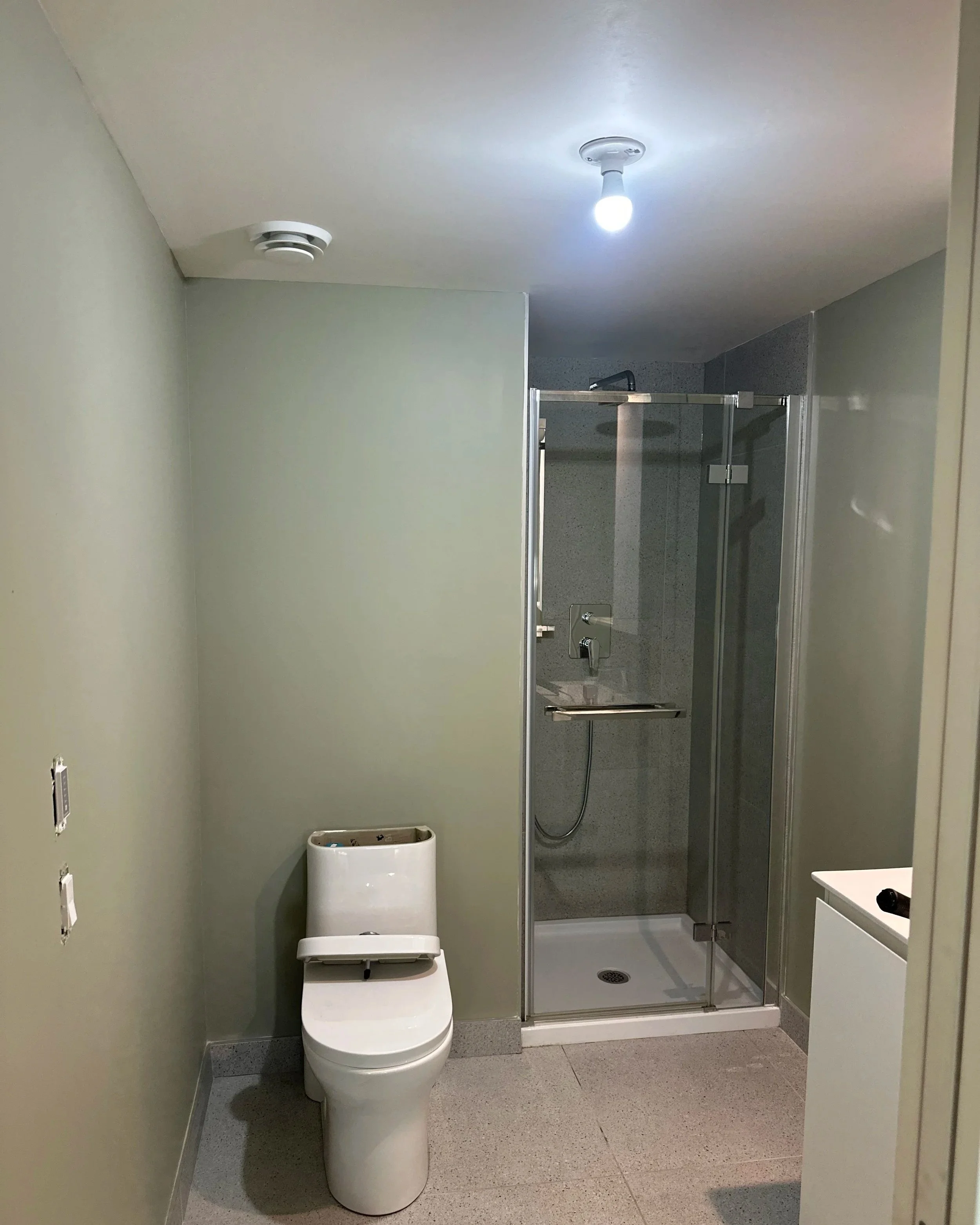 A partially renovated bathroom with a toilet, a glass shower enclosure, a small white sink, and unpainted wall outlets.