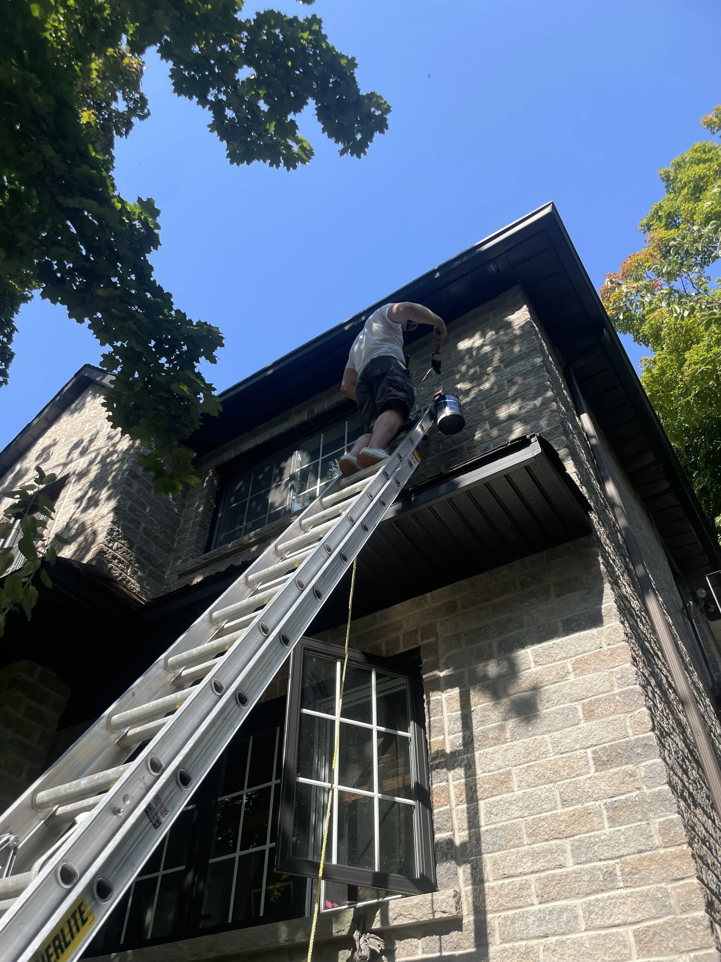 A person standing on a ladder outside a brick house painting or working near the roofline under a bright blue sky, surrounded by trees.