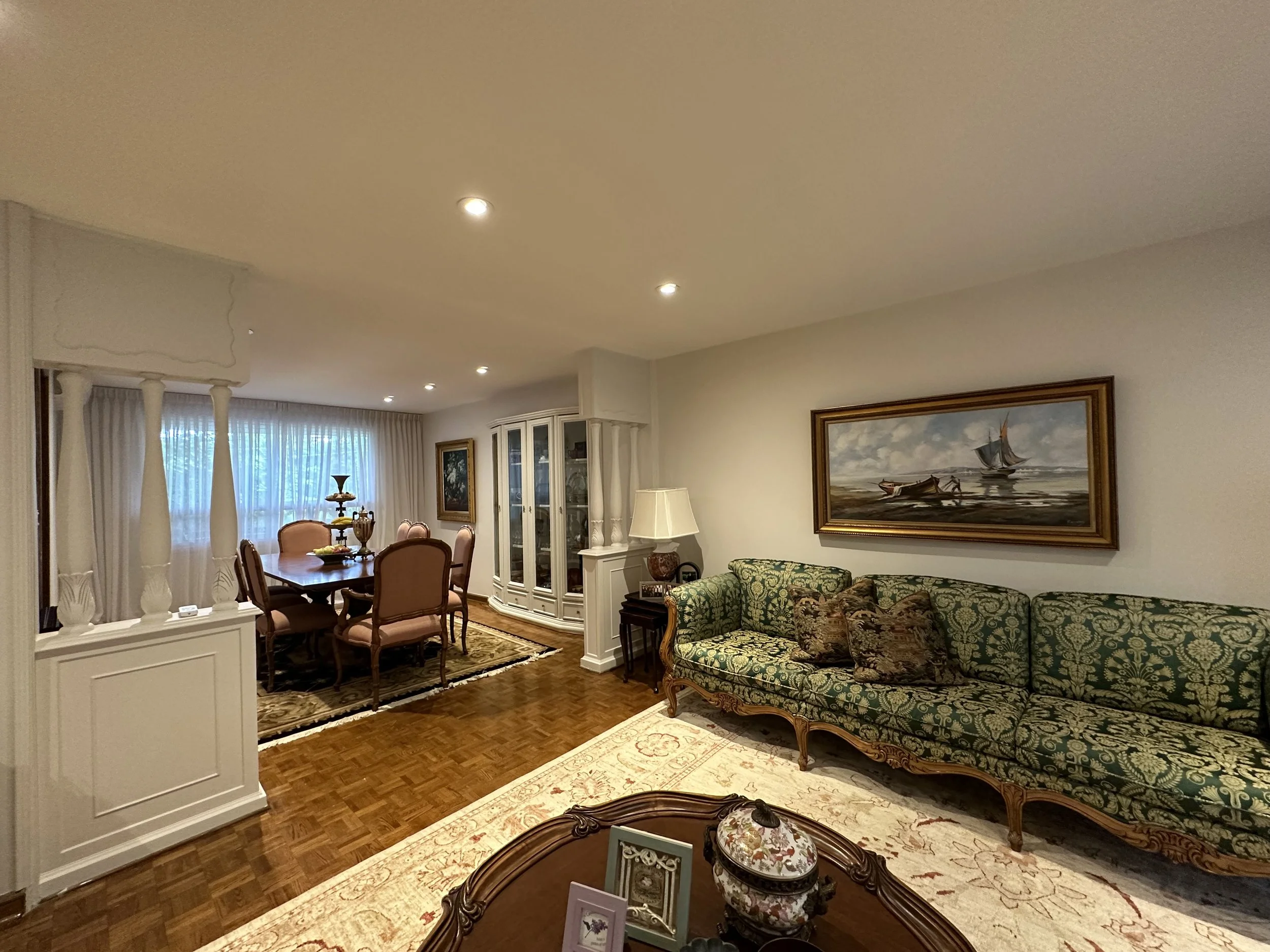 Interior of a living room featuring a dining table with chairs, a sofa with patterned upholstery, a framed painting of a boat on water, and a window with curtains.