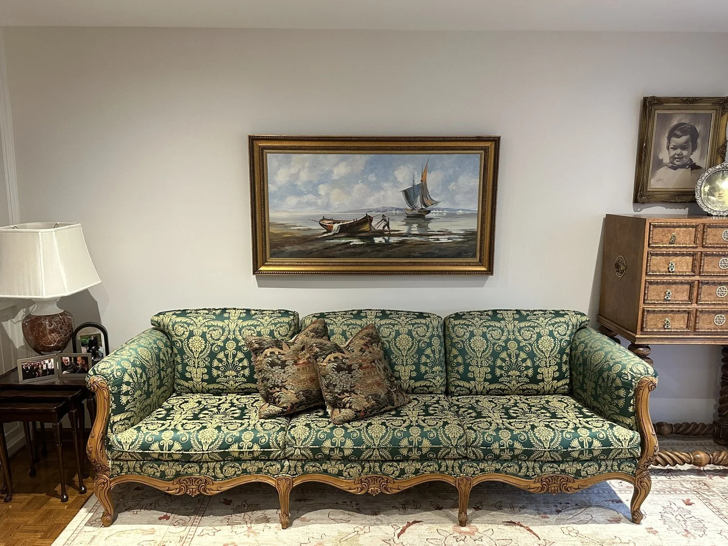 Vintage green and gold patterned sofa with three decorative pillows, a side table with picture frames and a lamp, a painting of boats on the water hanging on the wall, a wooden cabinet with drawers, a framed black and white photograph of a child, and a decorative plate, all in an elegant living room.